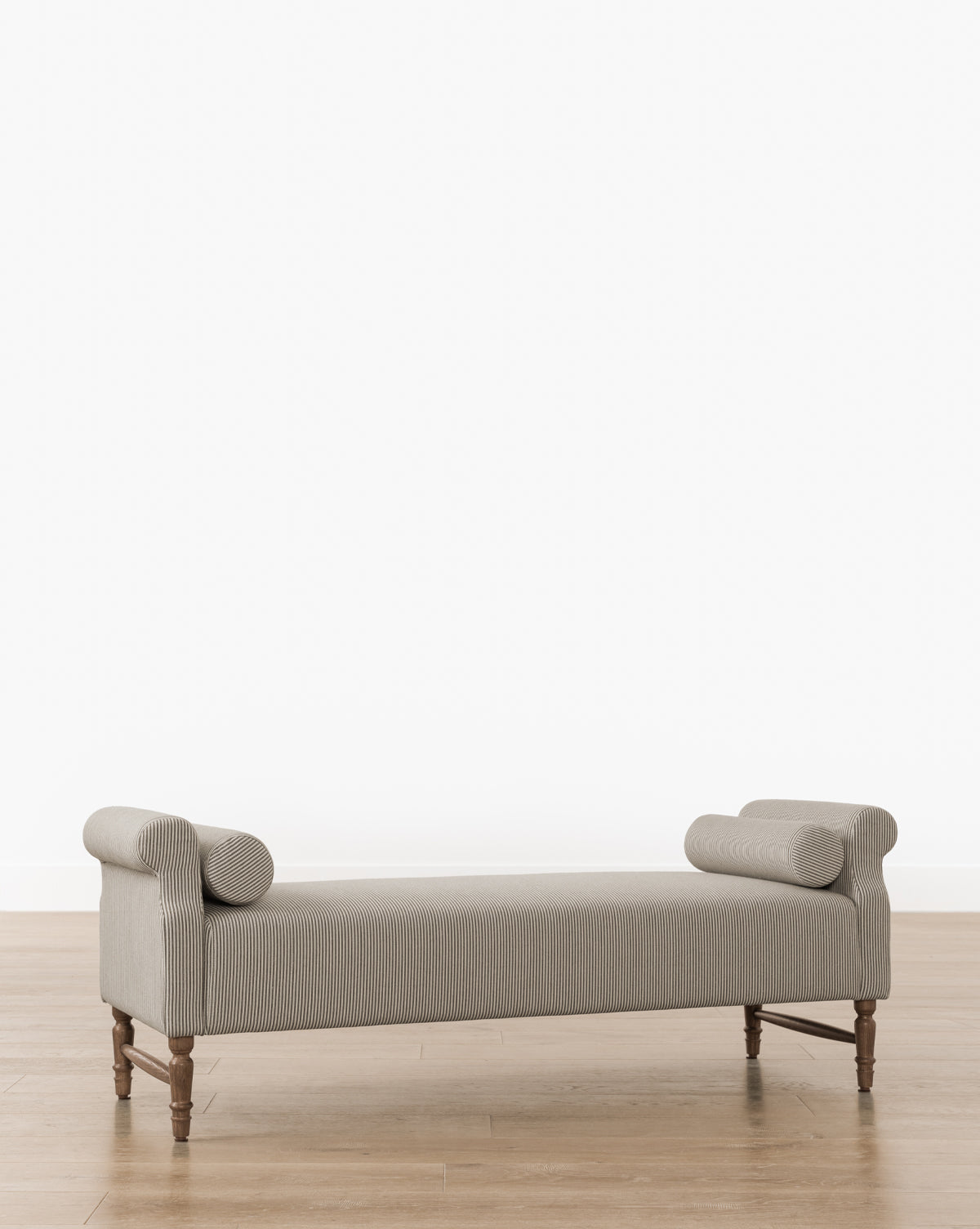 The Asher Bench showcases beige upholstered seating with striped linen and rounded bolster pillows at each end, supported by wooden legs, all set against a light wood floor and plain white background by McGee & Co.