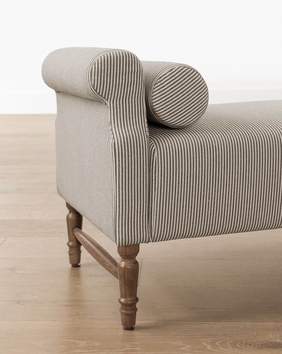 A close-up of the Asher Bench, featuring striped linen upholstery, rolled armrests, matching bolster pillows, and wooden legs set on a light wood floor, by McGee & Co.