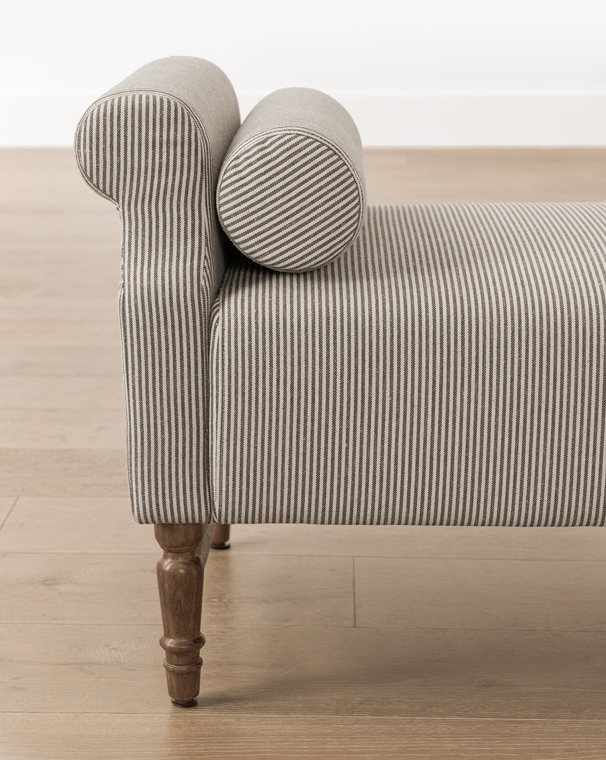 The Asher Bench, featuring gray and white striped linen, a rolled arm with bolster pillows on one end, and wooden legs, sits on a light wood floor against a white wall by McGee & Co.