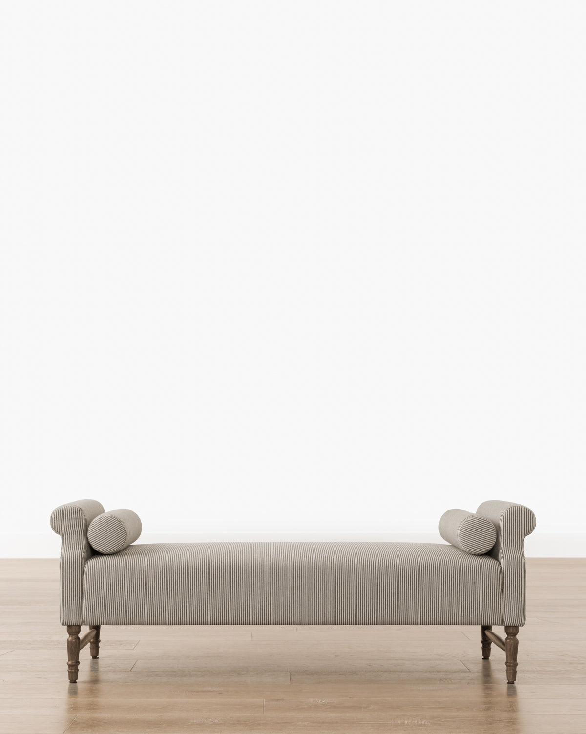 The Asher Bench offers gray upholstered seating with striped linen bolster pillows at each end, supported by wooden legs, and is shown on a wooden floor against a plain white background, by McGee & Co.