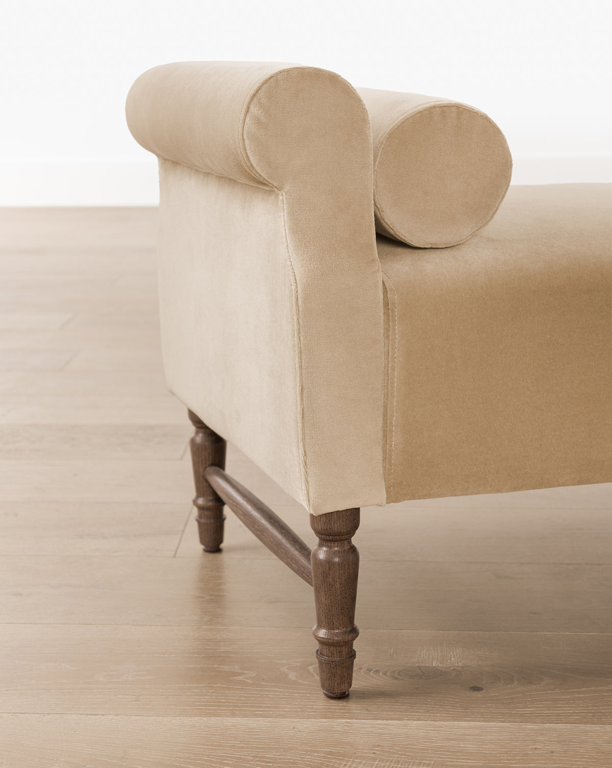 The Asher Bench is a beige upholstered chaise lounge with a rolled armrest, round cushion, bolster pillows, and wooden legs, shown on a light wooden floor, by McGee & Co.