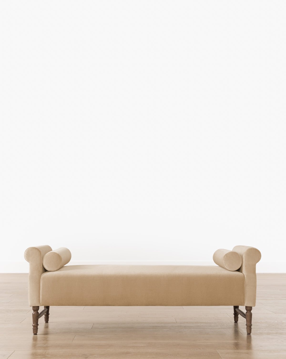 The Asher Bench, with a beige upholstered seat, rolled arms, and two bolster pillows, stands on wooden legs against a plain white backdrop by McGee & Co.