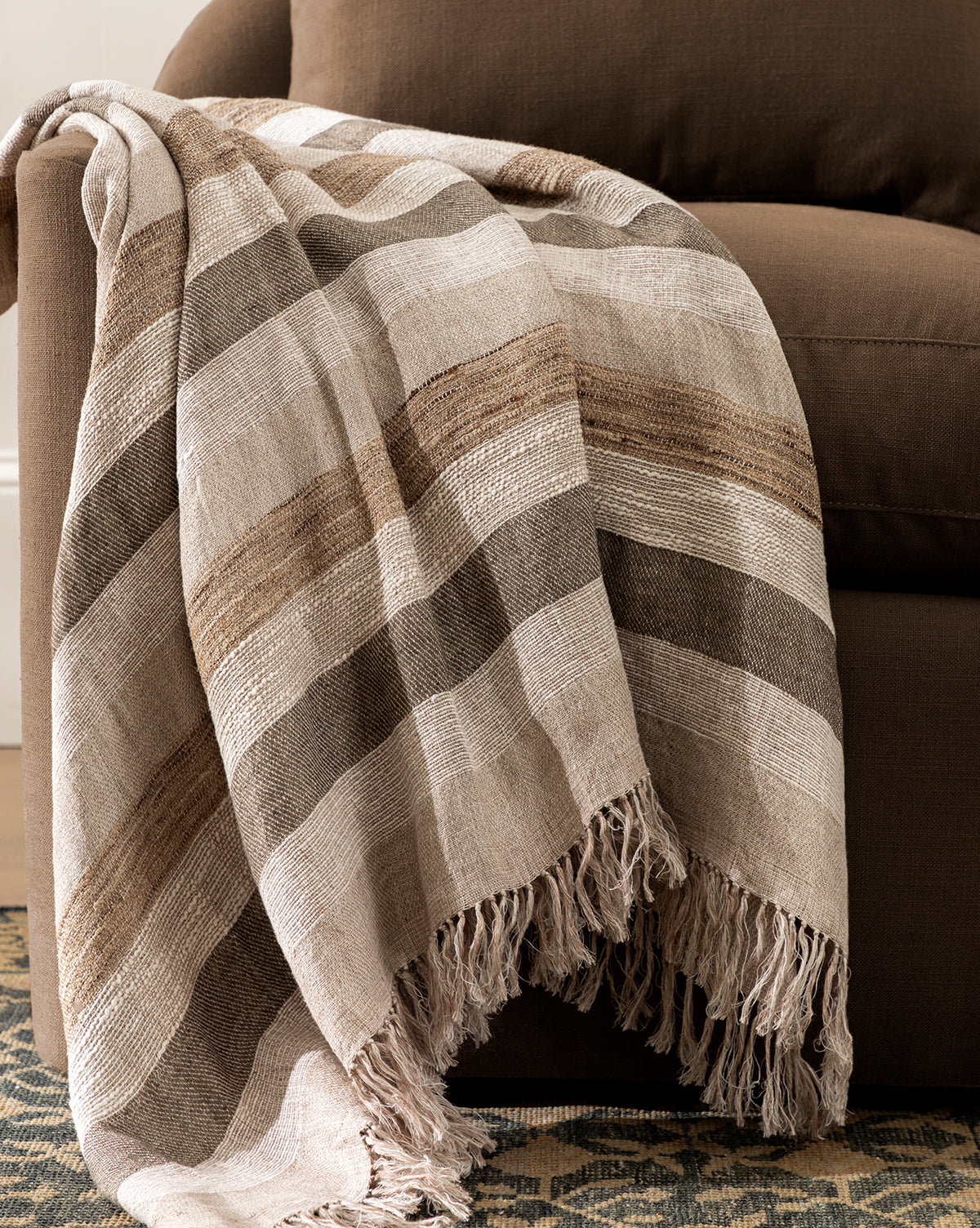 The Ashwood Throw, featuring brown, beige, and cream stripes and fringed edges, is draped over the arm of a brown upholstered sofa. A patterned rug can be seen on the floor nearby.