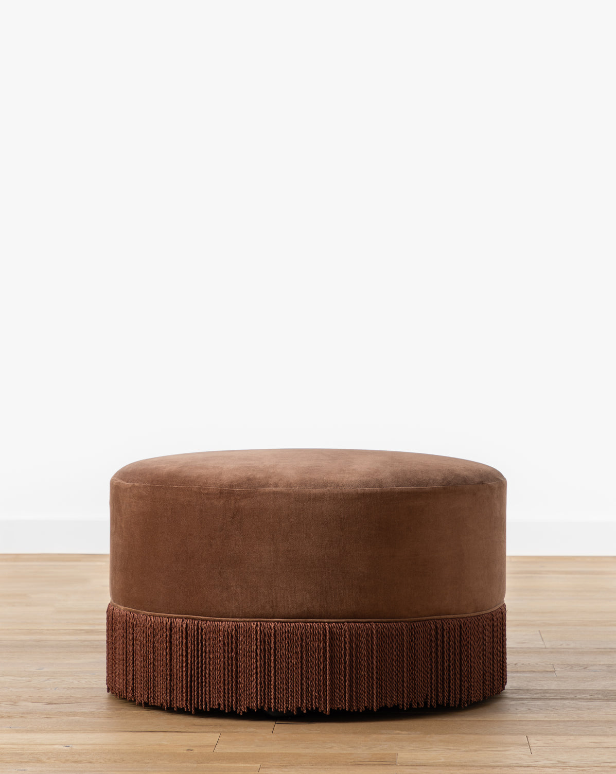 Certainly! Here’s your blended sentence with McGee & Co. at the end: The Makerspalm Astrid Fringe Ottoman, a round brown velvet piece with mid-century design and decorative fringe trim, sits on a light wood floor against a plain white wall, McGee & Co.