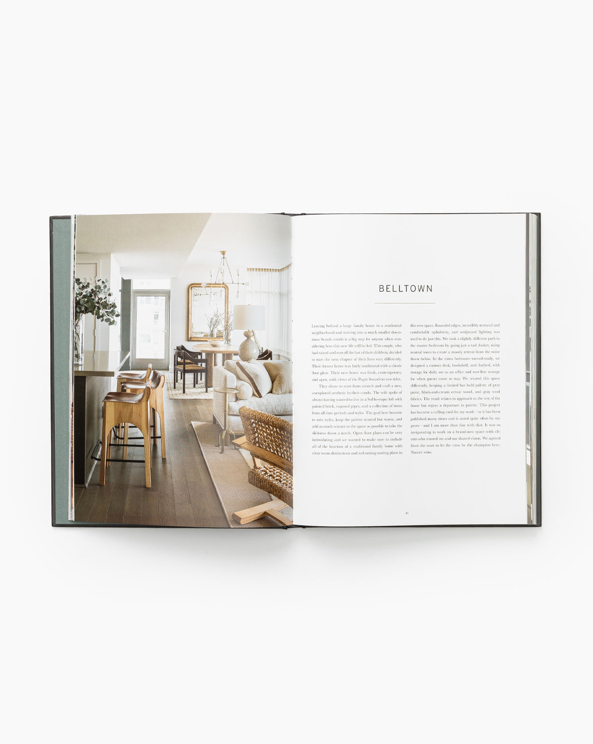 An open book titled At Home shows a photo of a modern, bright living and dining space inspired by Brian Paquette’s design on the left, with text titled Belltown on the right. The room features wooden furniture and neutral decor.