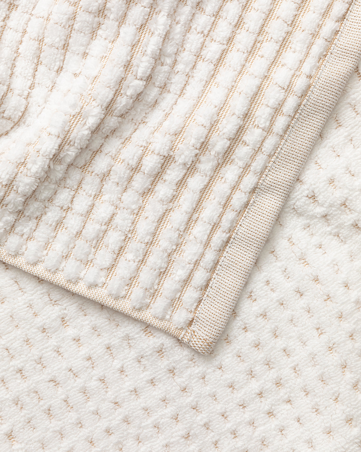 A close-up of a folded white waffle weave towel from our Atrani Tan Organic Bath Collection, featuring a textured striped pattern and beige trim, displayed on a subtly dotted white surface by McGee & Co.