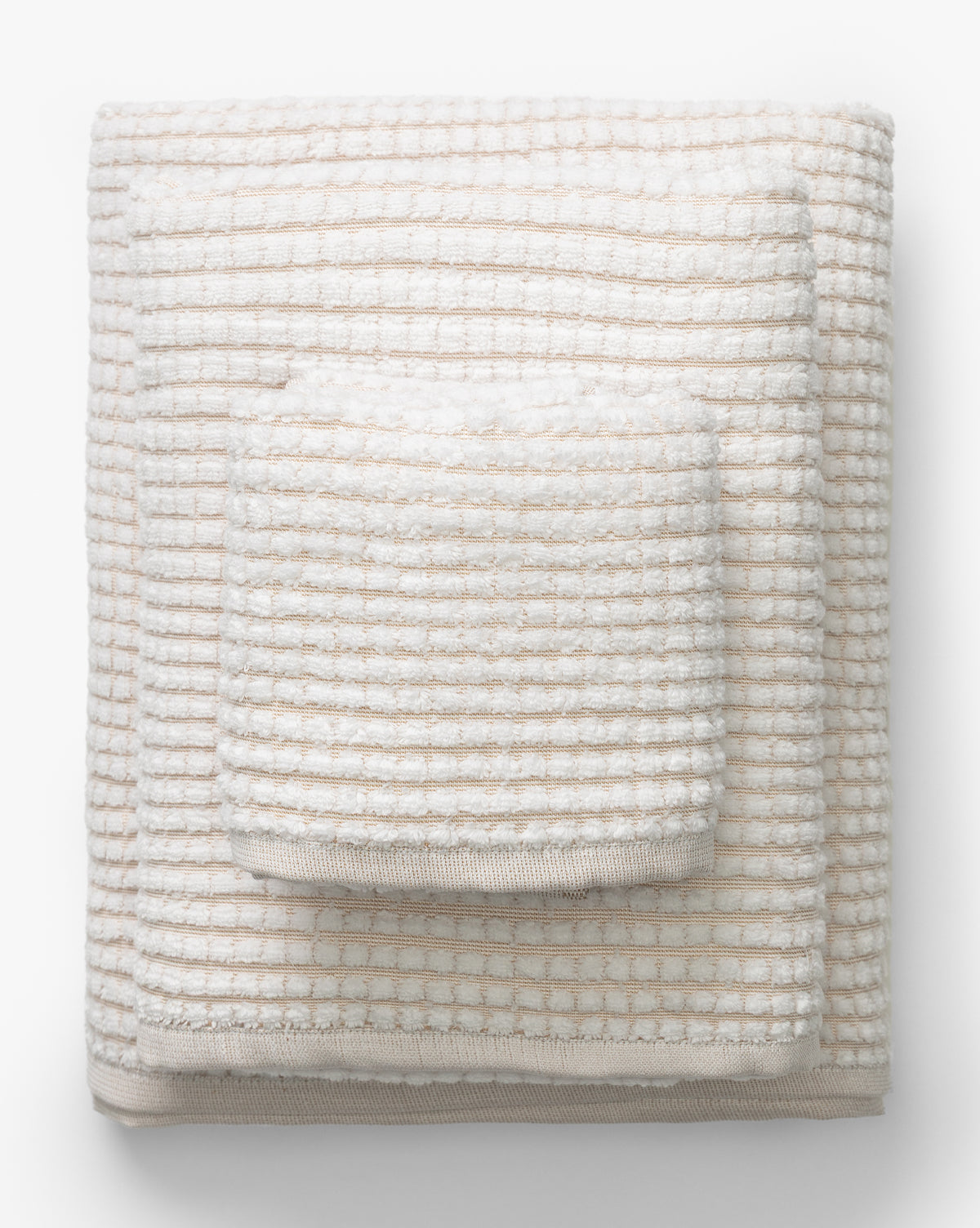 A neatly folded stack of three cream and beige striped towels in varying sizes from the Atrani Tan Organic Bath Collection, arranged largest to smallest on a white background, by McGee & Co.