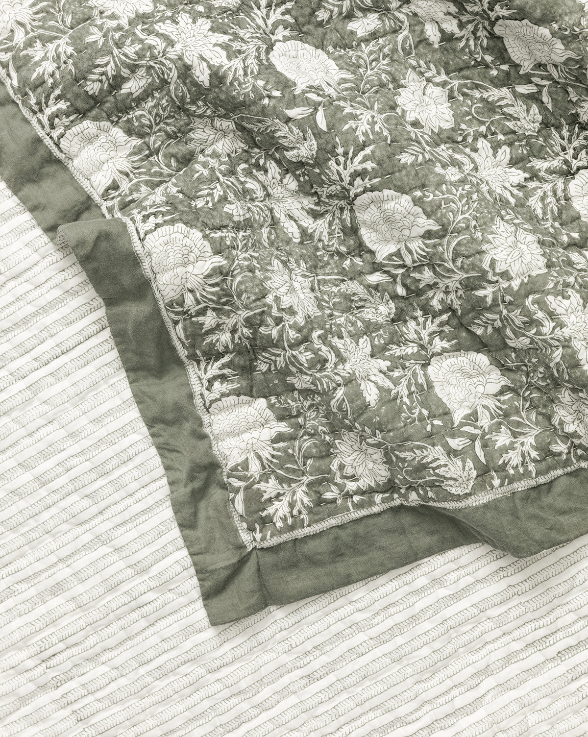 The Audrey Sage Block Print Quilt, featuring white floral embroidery and ruffle edging, is layered over a white textured blanket with subtle stripes. Its corner is folded over to showcase the quilts detailed stitching and pattern by McGee & Co.