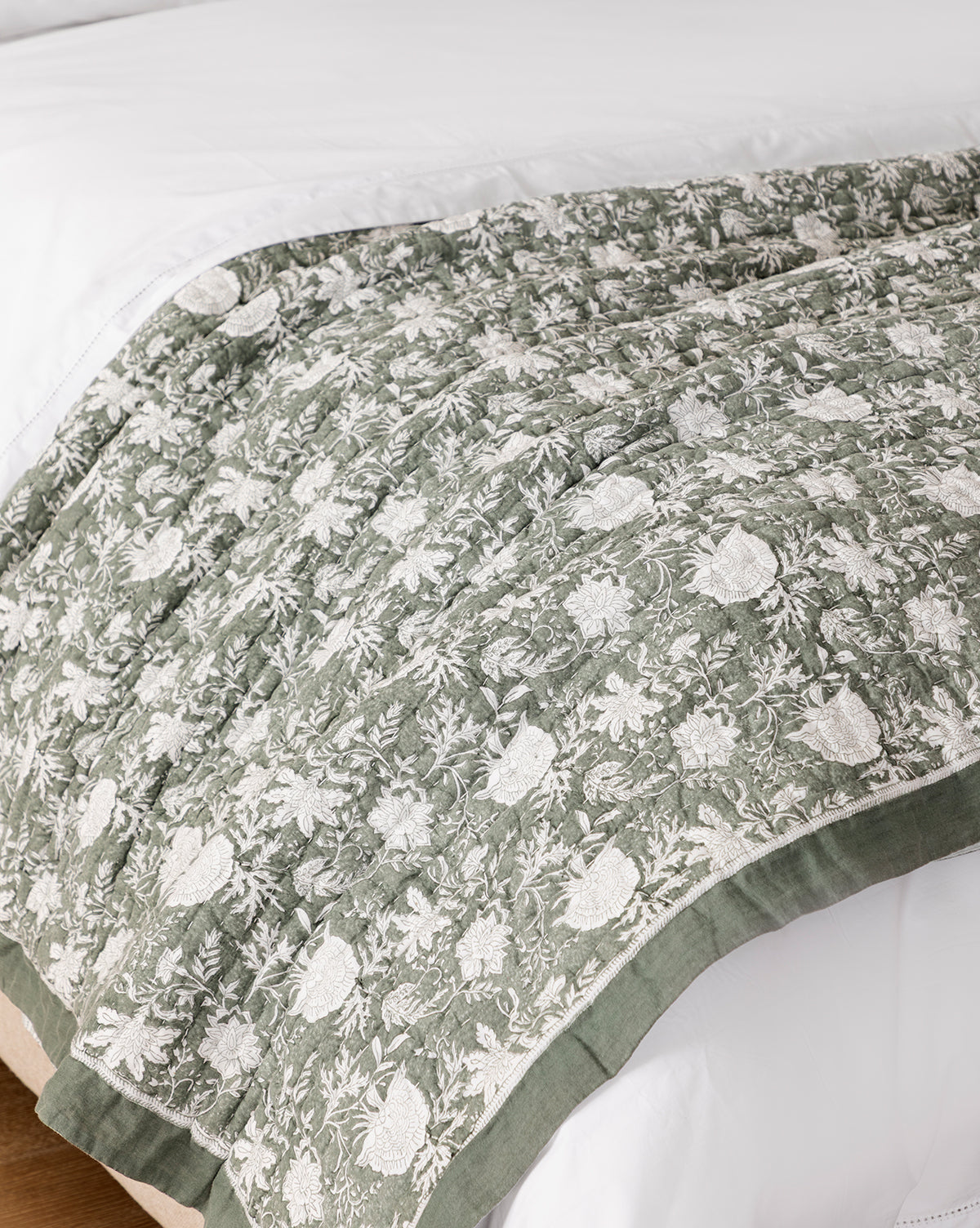 The Audrey Sage Block Print Quilt, featuring an intricate floral pattern and decorative border, is draped across the foot of a white bed, partially covering a white comforter and resting gracefully on a wooden floor, by McGee & Co.