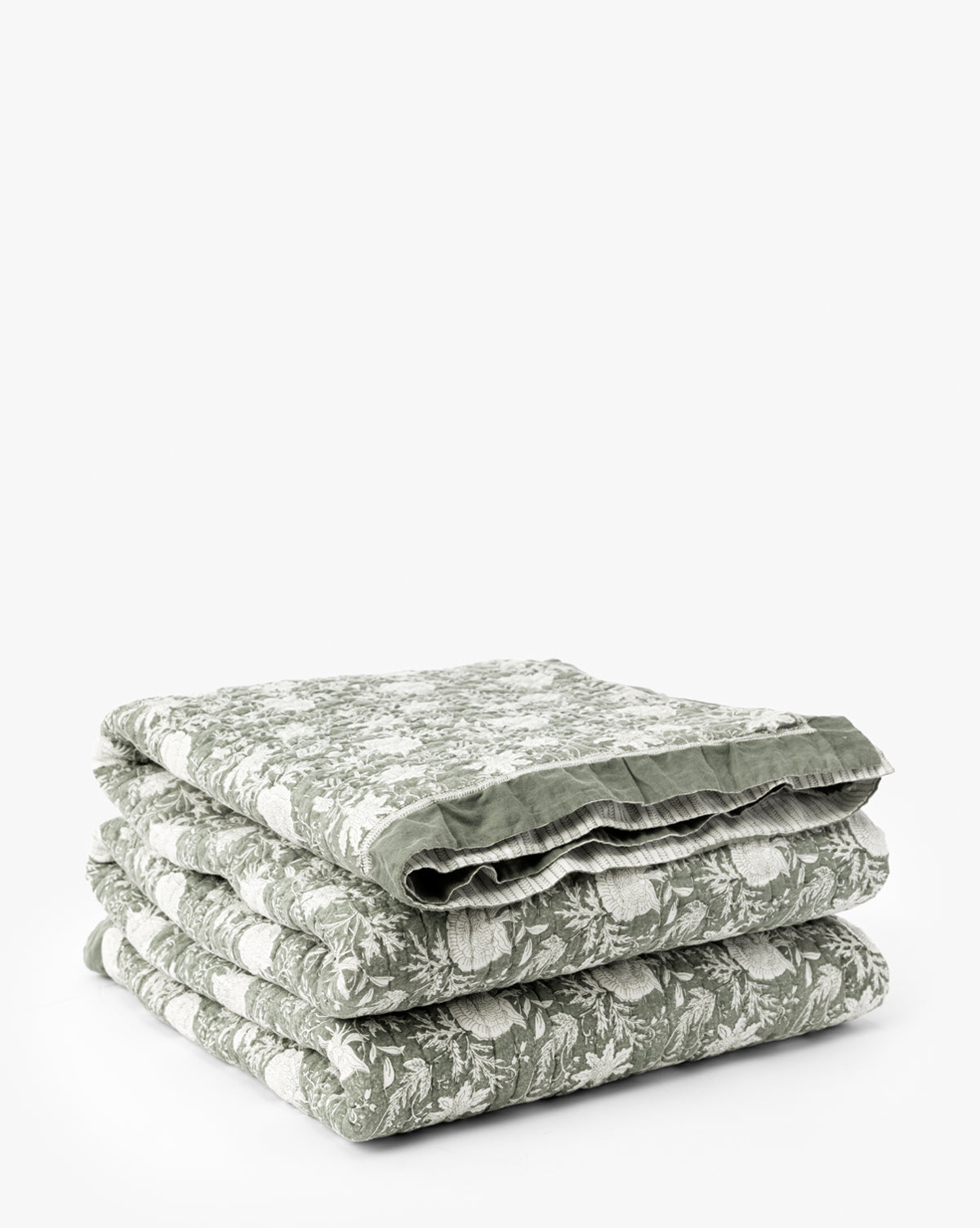 The Audrey Sage Block Print Quilt, featuring a delicate floral pattern and ruffle edging, is neatly folded and stacked on a plain white background by McGee & Co.