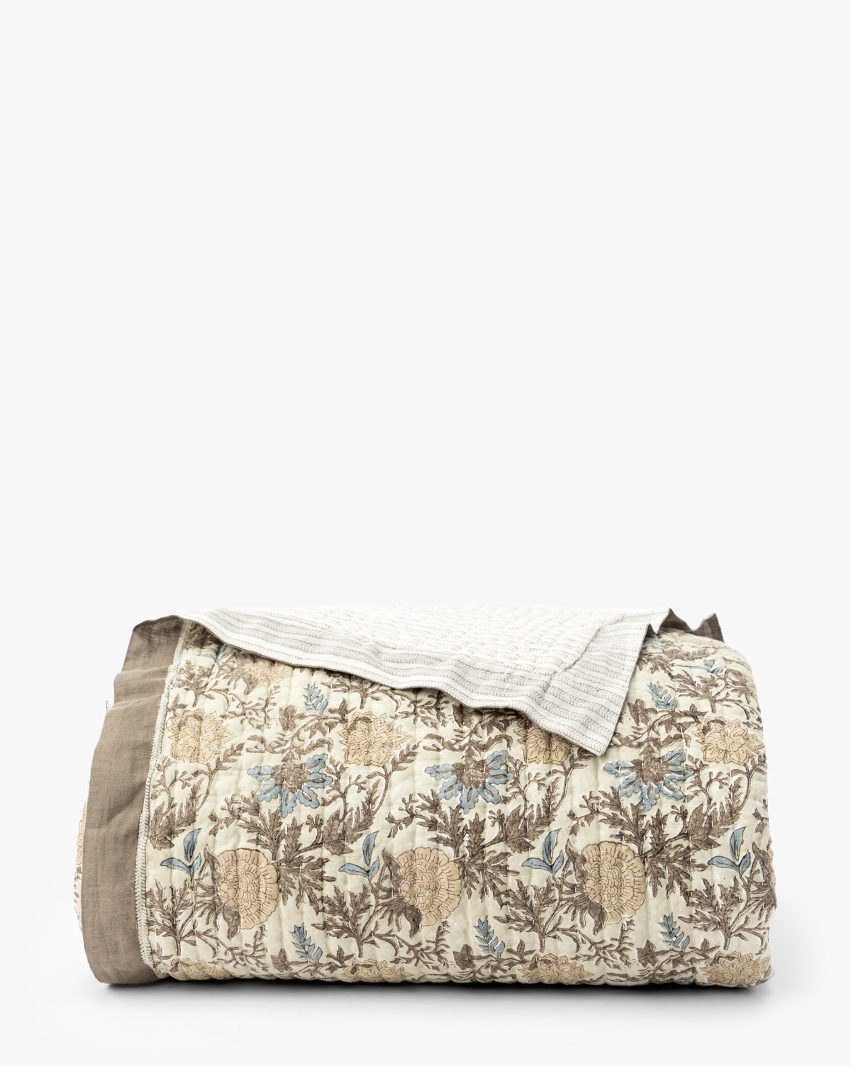 The Audrey Tan Block Print Quilt, folded to show its beige, brown, and blue floral leaf pattern with a solid taupe border and light beige textured corner, is displayed against a plain white background by McGee & Co.