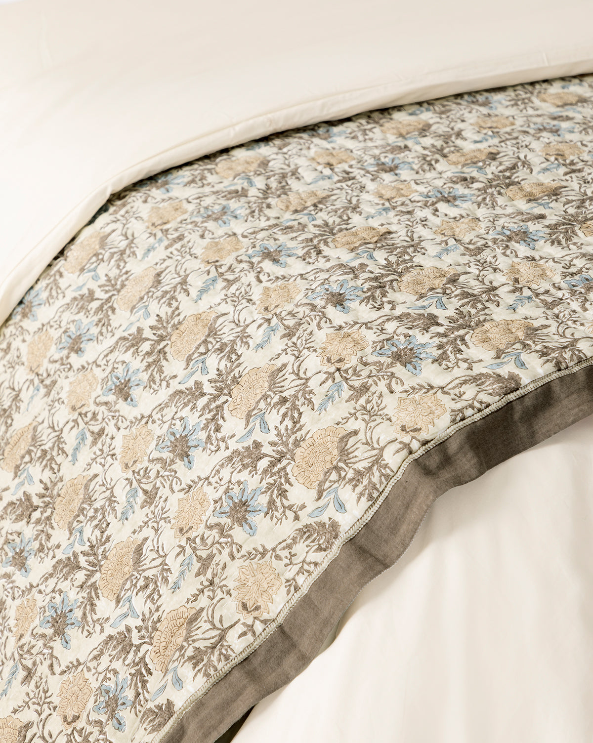 A close-up of a bed with the Audrey Tan Block Print Quilt, showcasing beige, brown, and light blue floral patterns on a white background with a gray border, layered over a plain white duvet, by McGee & Co.
