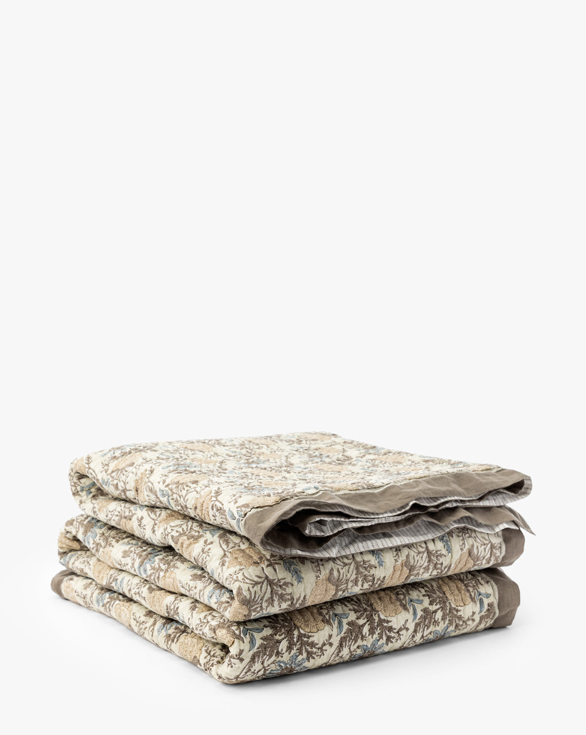 The Audrey Tan Block Print Quilt, featuring beige, brown, and blue floral patterns with delicate ruffle edging, is neatly folded and stacked against a plain white background by McGee & Co.