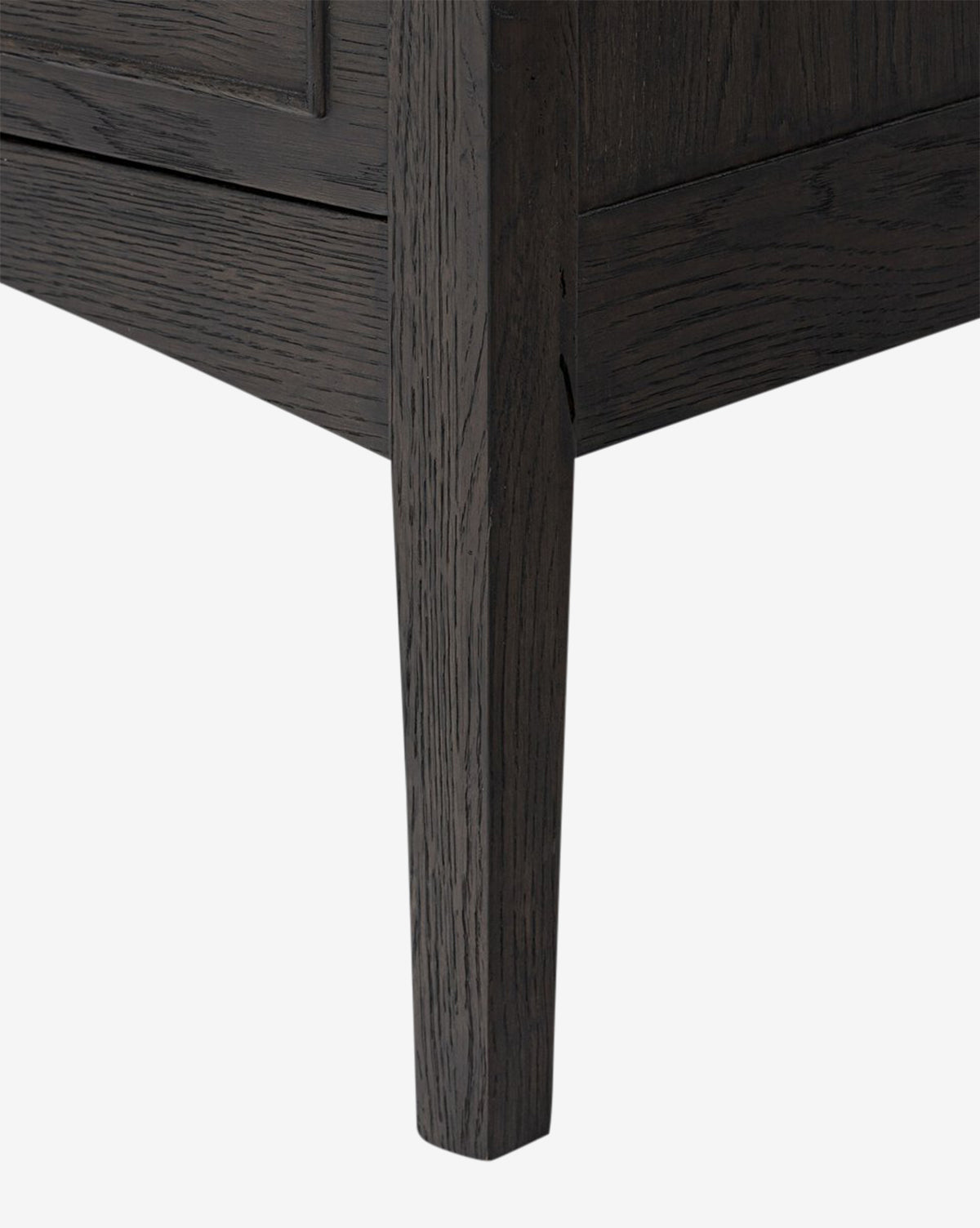 Close-up of the Aurelian Nightstands lower corner highlights its tapered leg and textured smoked oak grain—an elegant addition to any bedroom, photographed against a plain white background by McGee & Co.