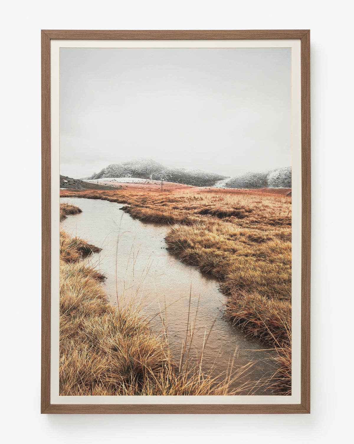 Autumn Marshes is a giclee print in a walnut wood frame, depicting a narrow stream winding through golden-brown autumn grass with distant snow-dusted hills beneath an overcast sky, by McGee & Co.