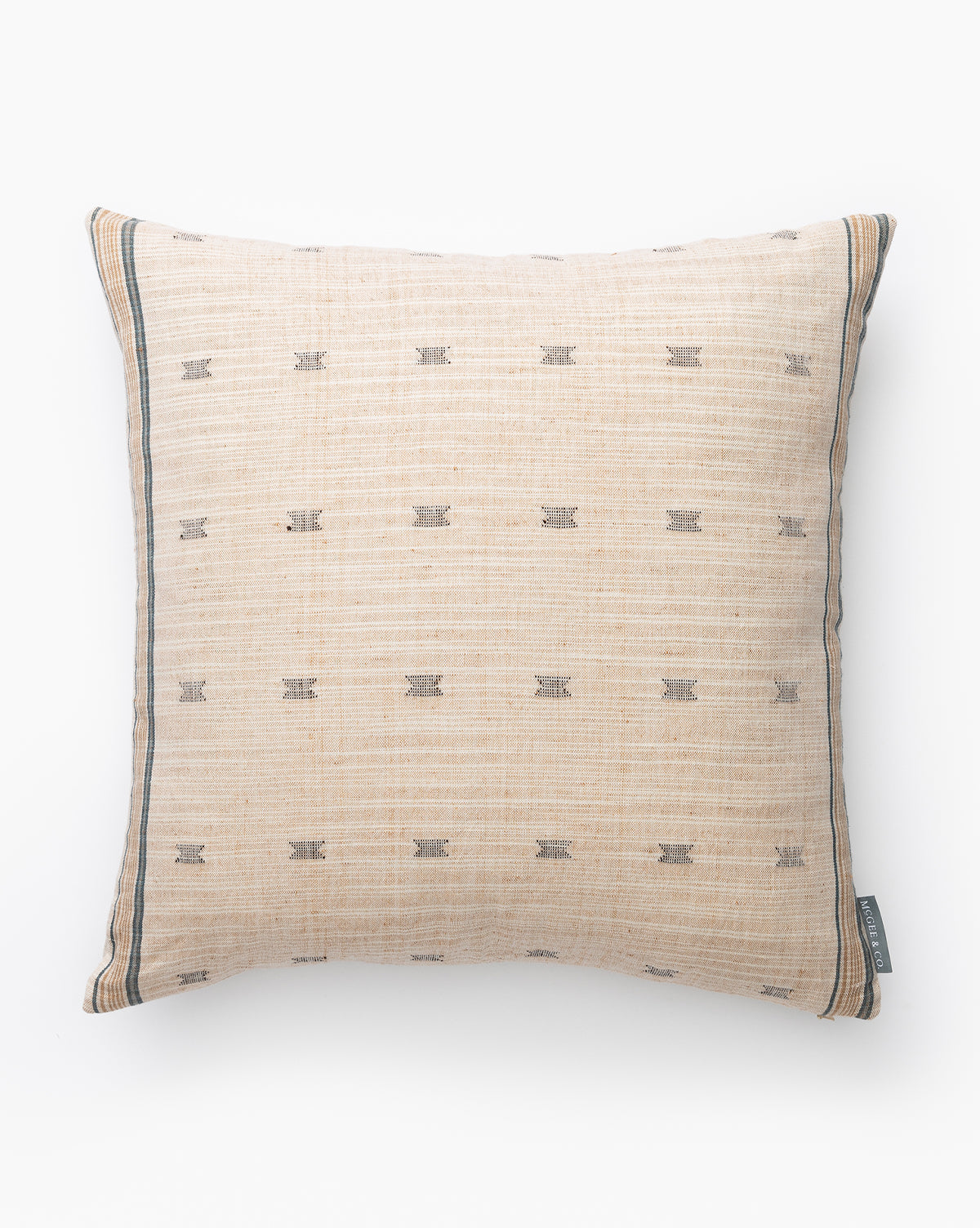 The Bali Pillow Cover is a square beige cotton pillow cover with subtle horizontal lines, small dark geometric patterns, thin dark borders at the edges, and a small fabric tag on one corner, shown against a white background by McGee & Co.