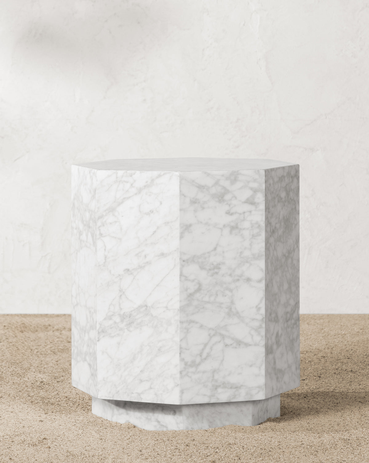 The Barcelo Outdoor Side Table, crafted from white marble with subtle grey veining, features a geometric design and stands on a sandy surface against a textured off-white wall, by McGee & Co.