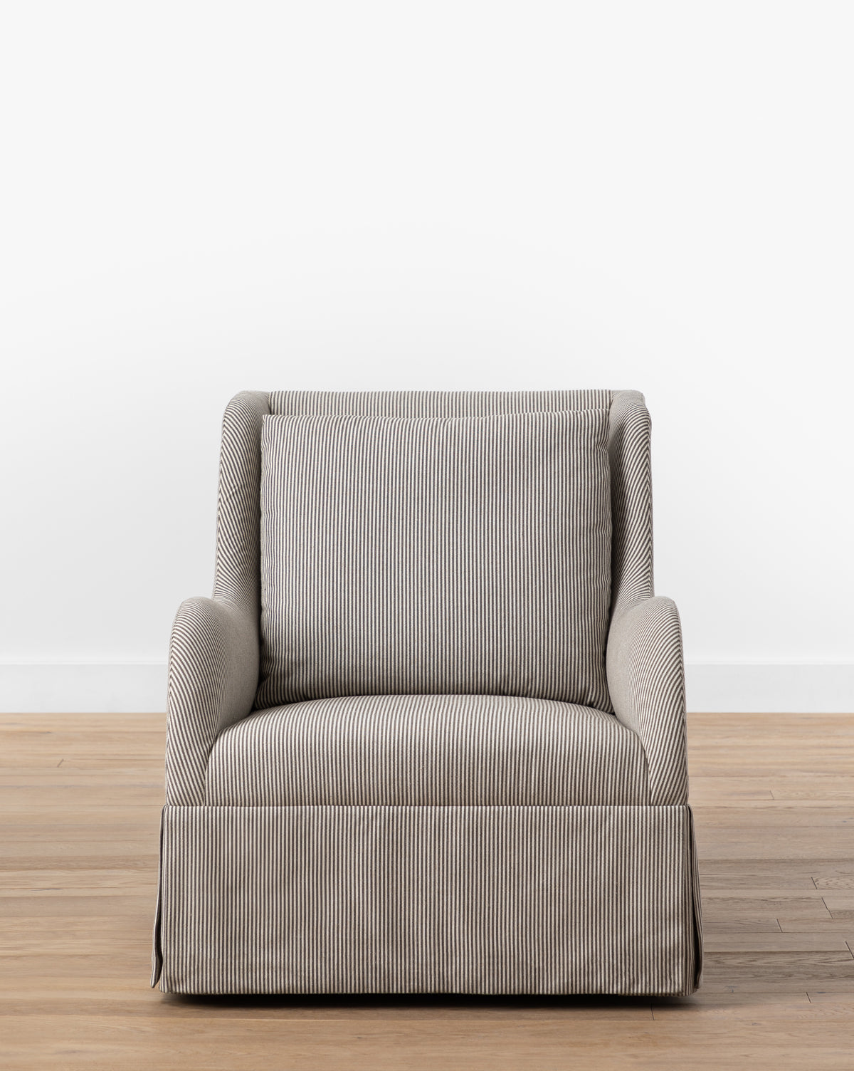 A gray and white striped Barnaby Glider with a skirted bottom is placed on a wooden floor by a plain white wall. Featuring a cushioned seat, backrest, and rounded armrests, the Barnaby Glider is perfect for any kids’ room, by McGee & Co.