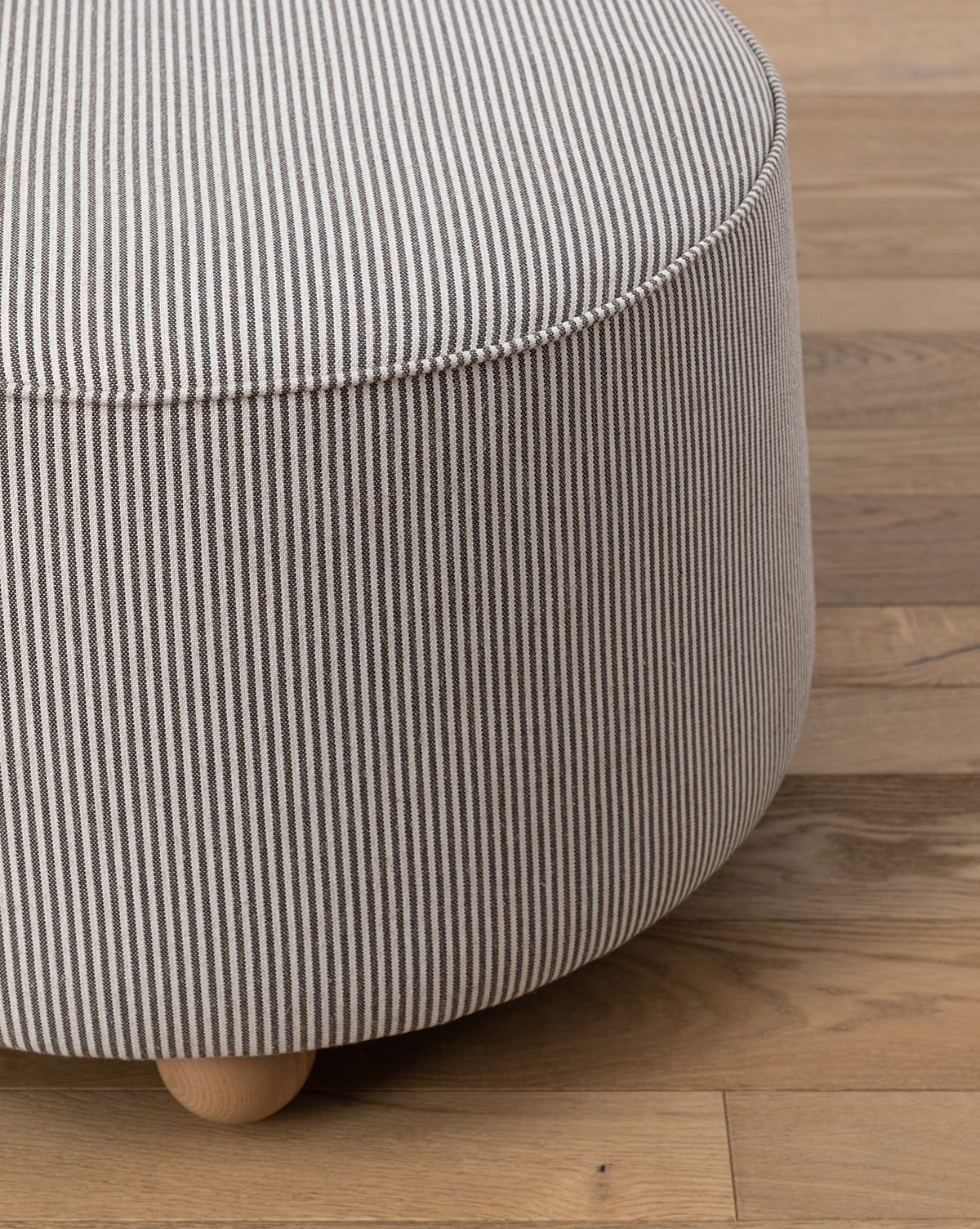 A close-up of the Barnaby Ottoman showcases its plush, round seat with black and white vertical striped fabric, resting on small ash wood feet atop a wooden floor, by McGee & Co.