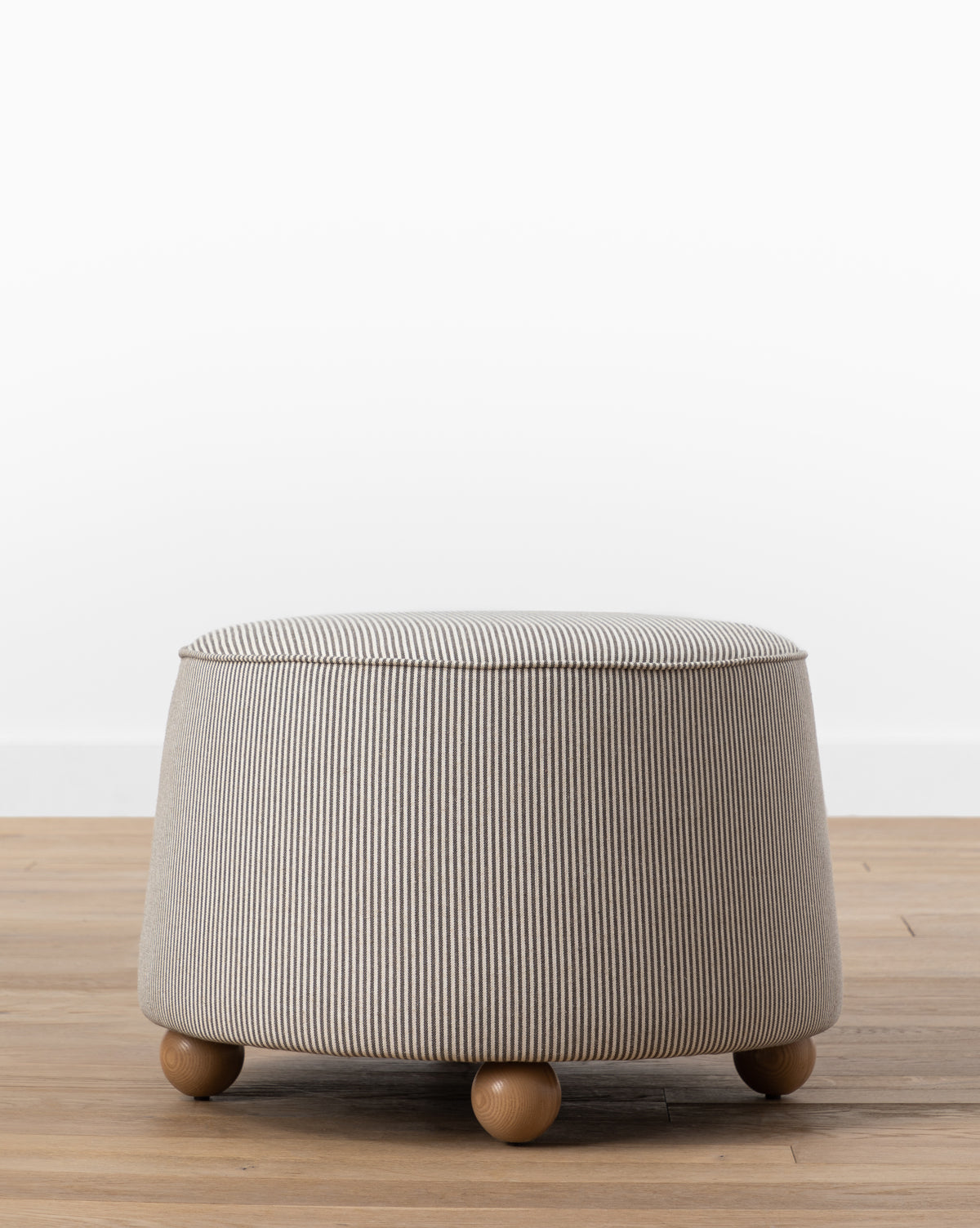 The Barnaby Ottoman is a round, small ottoman in light gray with subtle vertical stripes, featuring four short ash wood legs. It’s shown on a wooden floor against a white wall by McGee & Co.