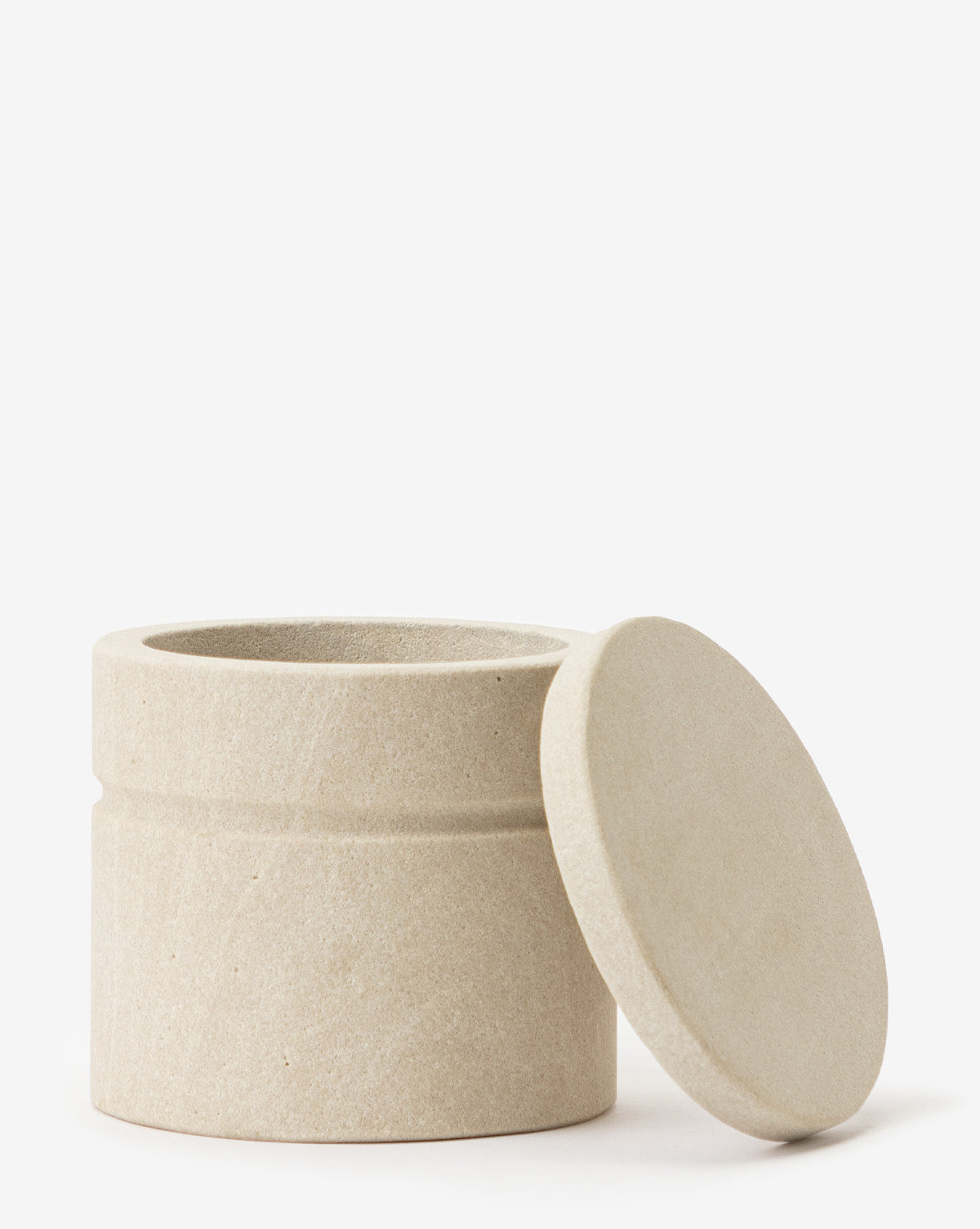The Barton Sandstone Canister is a round, beige container with a matching lid, shown open and leaning against its side. Its smooth design makes it perfect for bathroom countertops. Set against a white background.