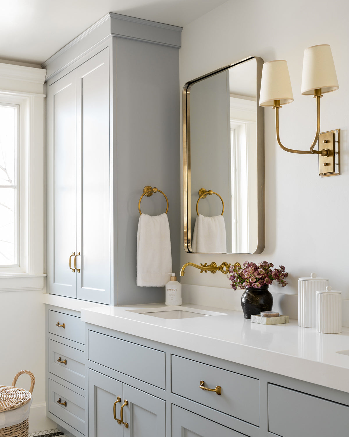 A modern bathroom with light gray cabinets, gold hardware and faucet, a white countertop, a rectangular gold-framed mirror, white towel, sconce, purple flowers in a vase, and the Fluted Lidded Bathroom Canister as an elegant accessory.