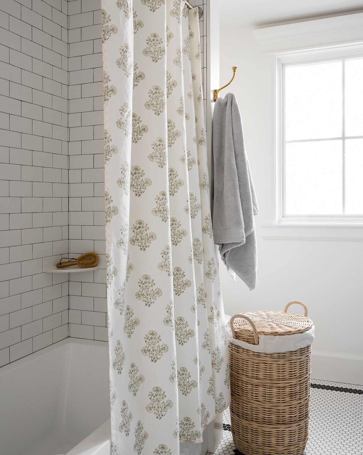 A bathroom featuring white subway tile, a Block Print Shower Curtain, a gold showerhead, a towel on a gold hook, and a lidded wicker laundry basket on black-and-white tile near a bright window.