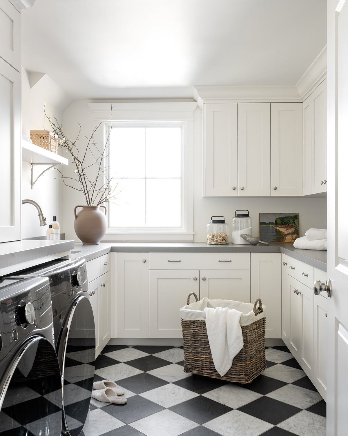 Bright laundry room with white cabinets, black and white checkered floor, washer and dryer, a Kobo Wheeled Basket holding white towels, natural light from a window, and a vase with branches decorating the counter.