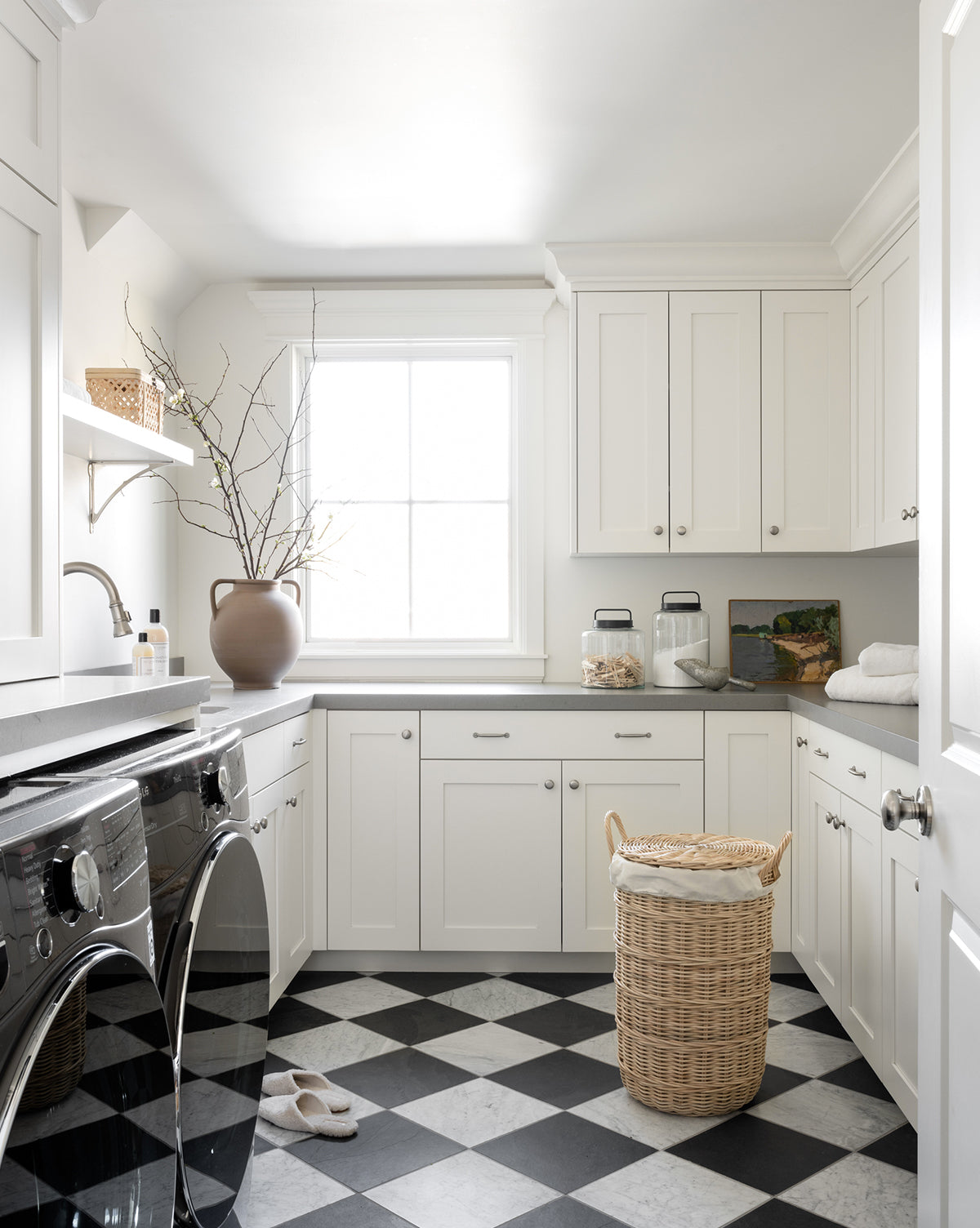 A bright laundry room features white cabinets, a black-and-white checkered floor, a washer and dryer, the Rattan Lidded Laundry Bin, a vase with branches, and sunlight streaming in through the window.