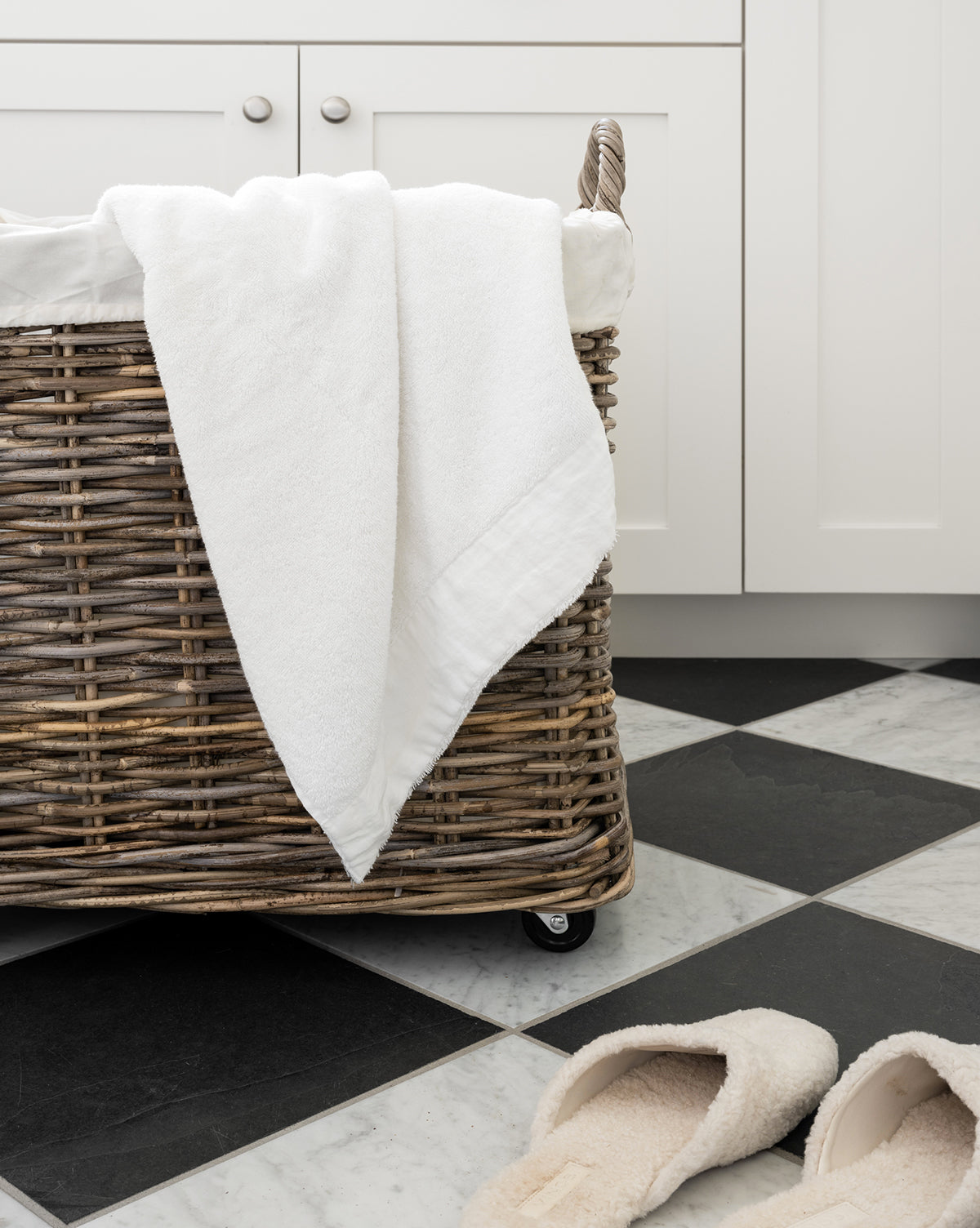 The Kobo Wheeled Basket with a white towel draped over it rests on a black-and-white checkered tile floor beside beige slippers in a bright, modern room featuring white cabinets.