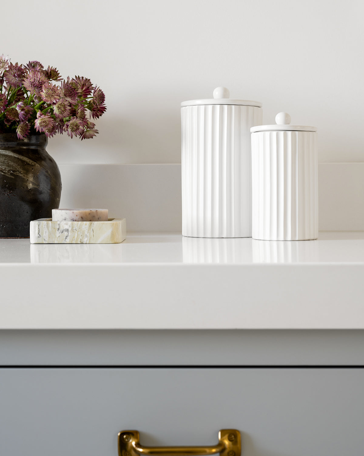 A minimalist countertop features two white Fluted Lidded Bathroom Canisters, a dark vase with purple flowers, and stone coasters, against a white wall above a gray drawer with a gold handle.