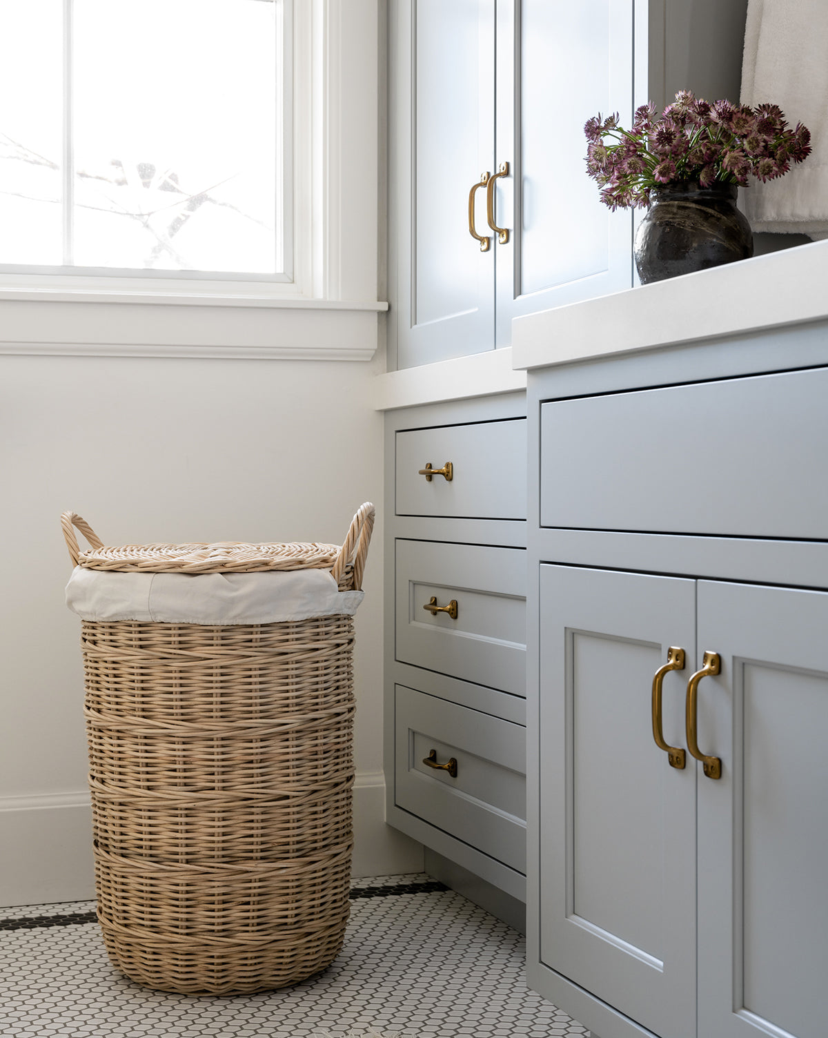 The Rattan Lidded Laundry Bin with handles sits next to light gray cabinets with gold handles in a bright laundry room. A vase of flowers decorates the countertop beneath a window streaming in natural light.