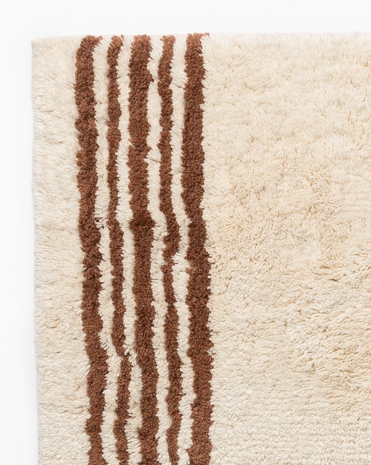 A close-up of the Beata Bath Mat, featuring a cream color with a textured surface and vertical brown stripes along one edge, displayed on a white background.