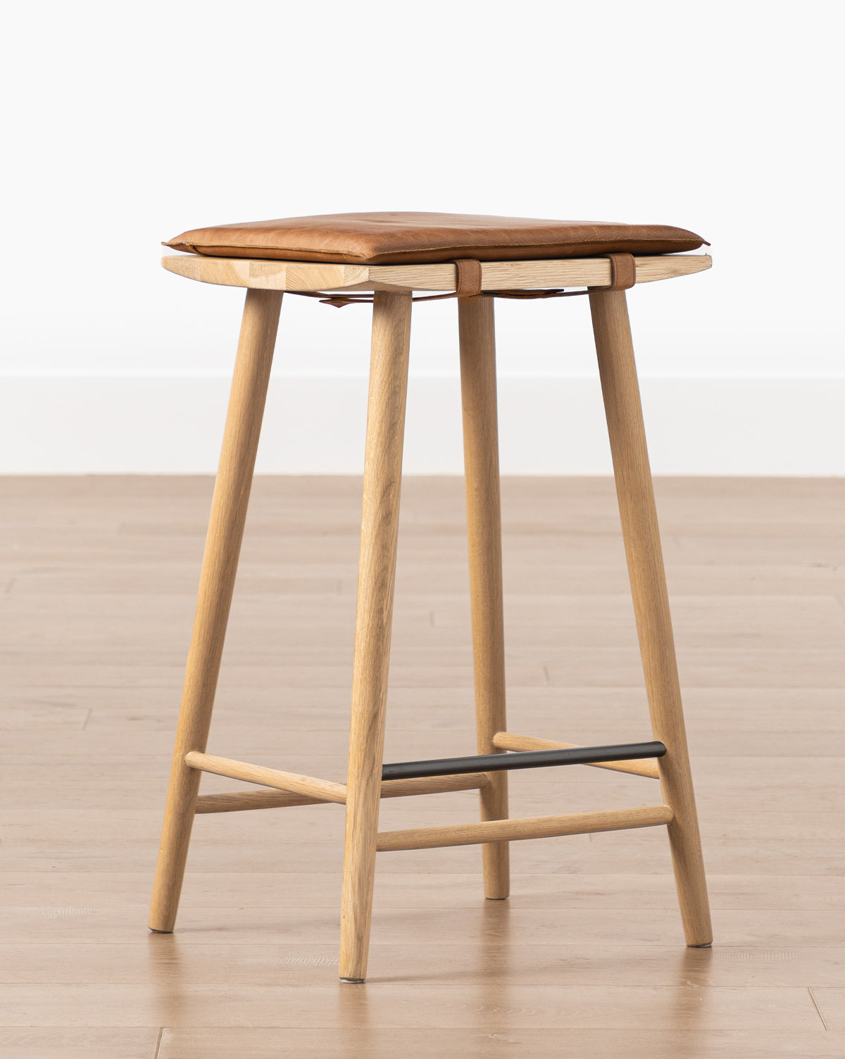 The Beau Counter Stool features a modern design with a wooden frame, four angled legs, a square brown leather seat cushion, and a black metal footrest, by McGee & Co.