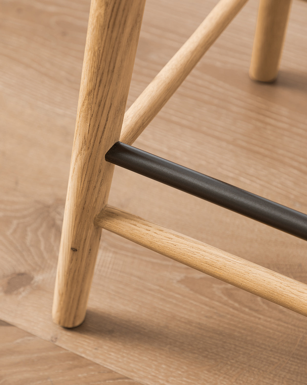 Close-up of the Beau Counter Stool’s wooden legs on a light wood floor, highlighting the sleek leather seat, black support bar, and matching wood bar—details that define this modern stool’s elegant design, by McGee & Co.