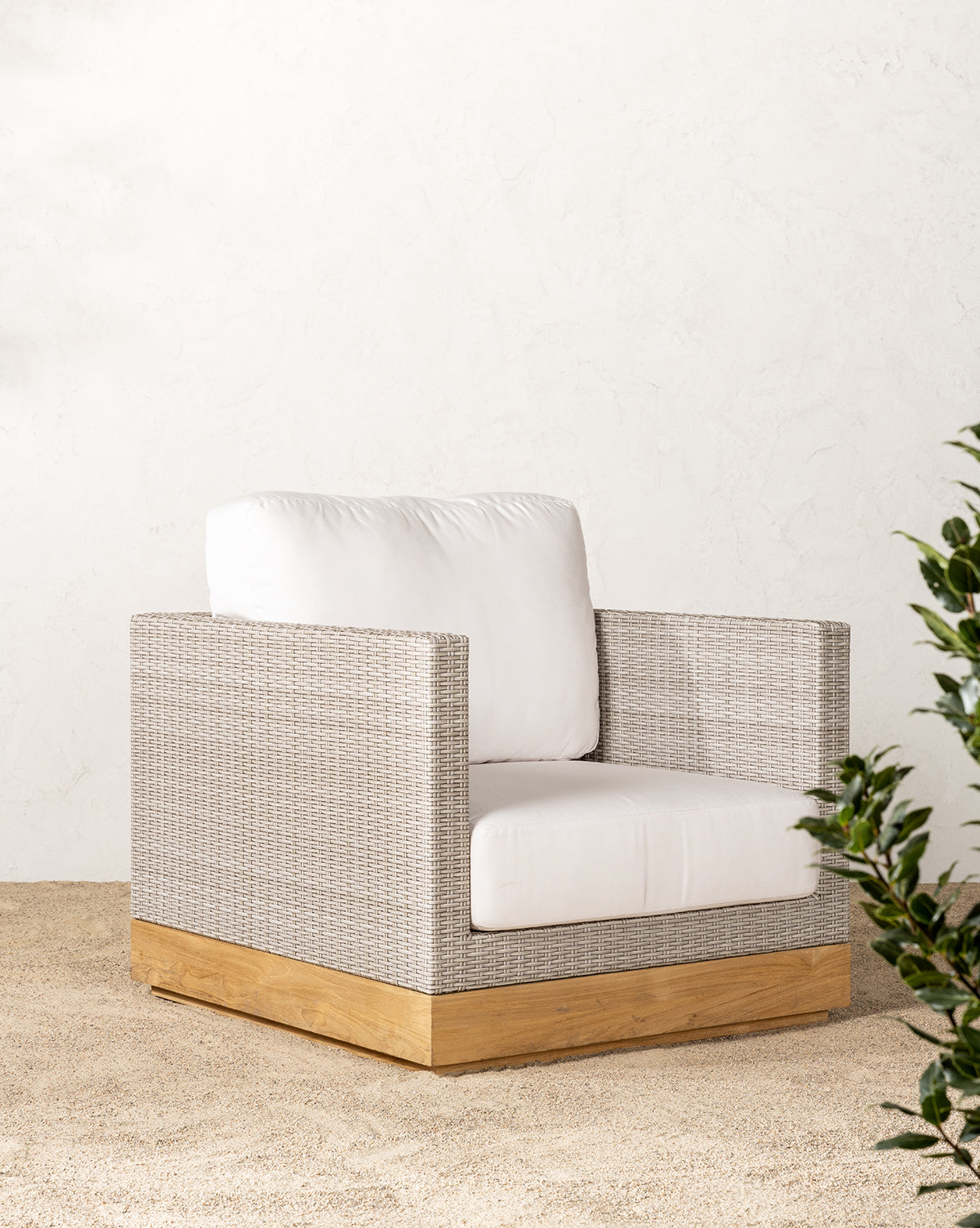 The Berdine Wicker Outdoor Chair, featuring a teak base, white cushions, and wicker exterior, is placed on sand near a light textured wall with green leafy plants partly visible on the right.