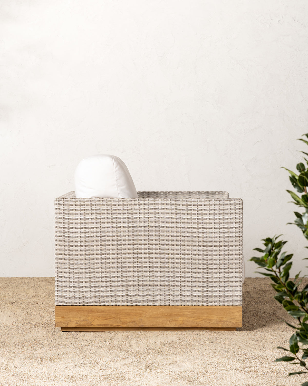 The Berdine Wicker Outdoor Chair, featuring a light woven texture, teak base, and white cushion, sits on sand against a white textured wall with green foliage in the foreground.