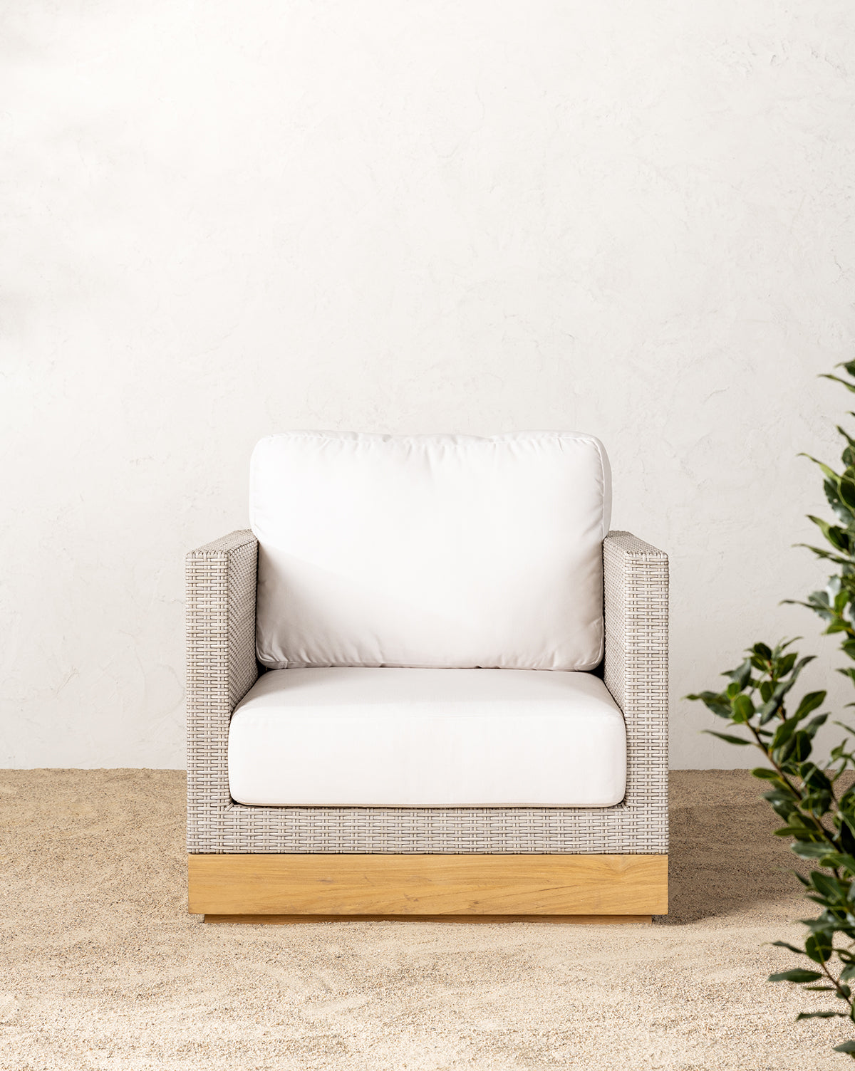The Berdine Wicker Outdoor Chair features a light woven frame, white cushions, and a teak base. It sits on sand against a textured white wall, with green leaves visible in the lower right corner.