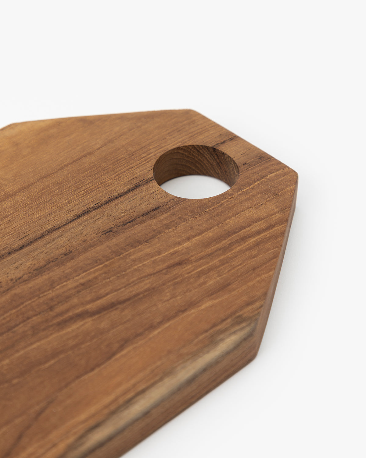 A close-up of the Bevelled Edge Cutting Board, pentagon-shaped with a round hole near one corner, crafted from teak root and set against a white background—a must-have for any home chef, by McGee & Co.