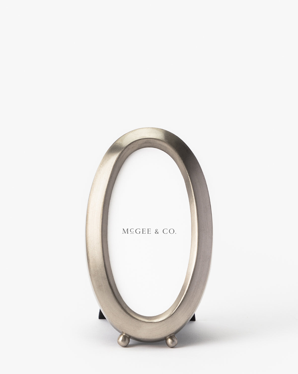 The Blanche Pewter Picture Frame features a sleek oval silver design with a modern, sophisticated style. Standing upright on two small round feet, it displays a MEGEE & CO. placeholder card against a plain white background.