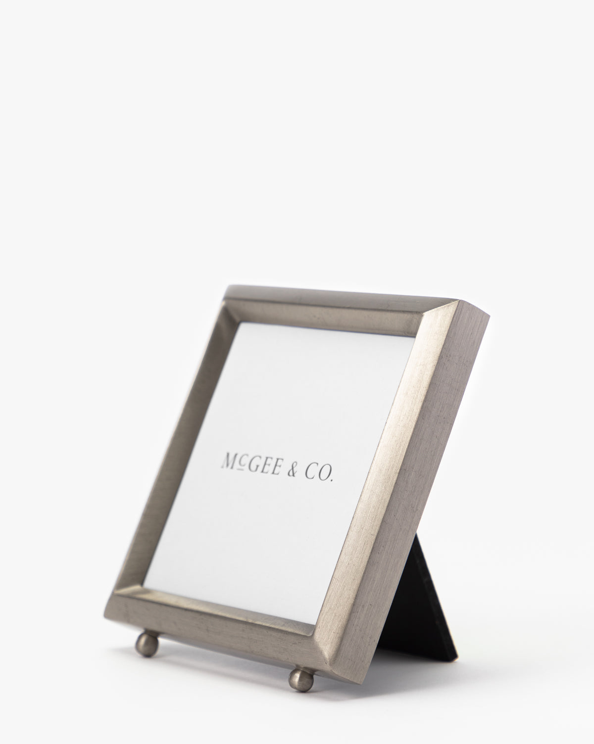 The Blanche Pewter Picture Frame, a square metallic design with iron corners and stand, holds an MCGEE & CO. insert and exudes sophistication as it stands angled against a plain white background.