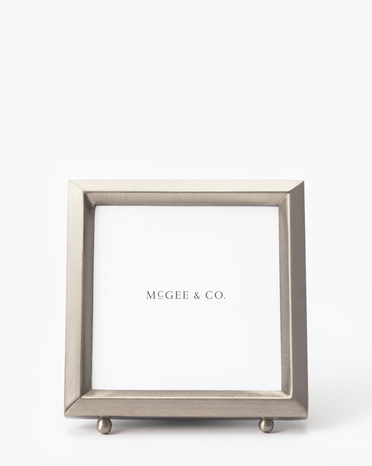 The Blanche Pewter Picture Frame is a square, silver metal frame with a simple modern design, iron corners, and two small round feet. It features a white placeholder displaying McGee & Co.