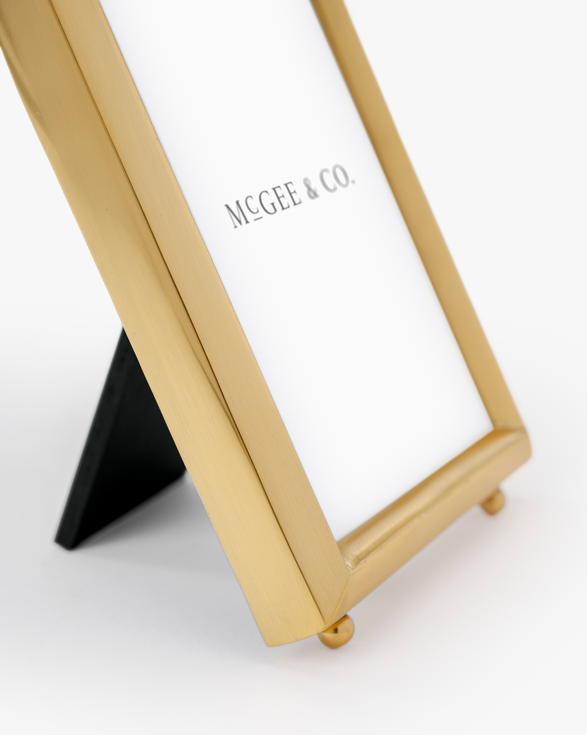 A close-up of the Blanche Picture Frame in polished brass stands on a black support, displaying "McGee & Co." on the paper inside, set against a plain white background, by McGee & Co.