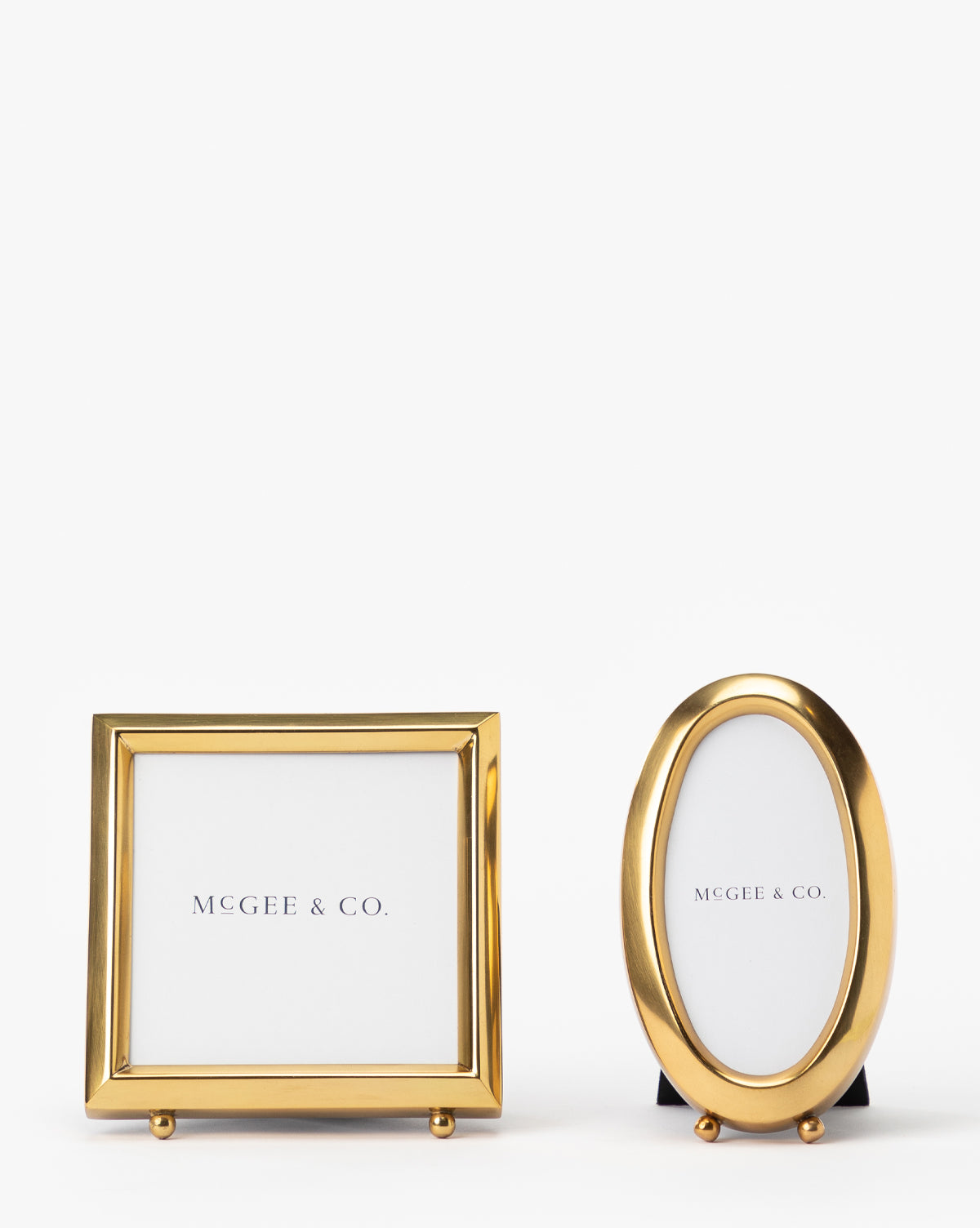 Two small Blanche Picture Frames, one square and one oval, feature an antique brass finish and white McGee & Co. inserts. These gold metal frames stand upright on a plain white background, adding vintage charm.