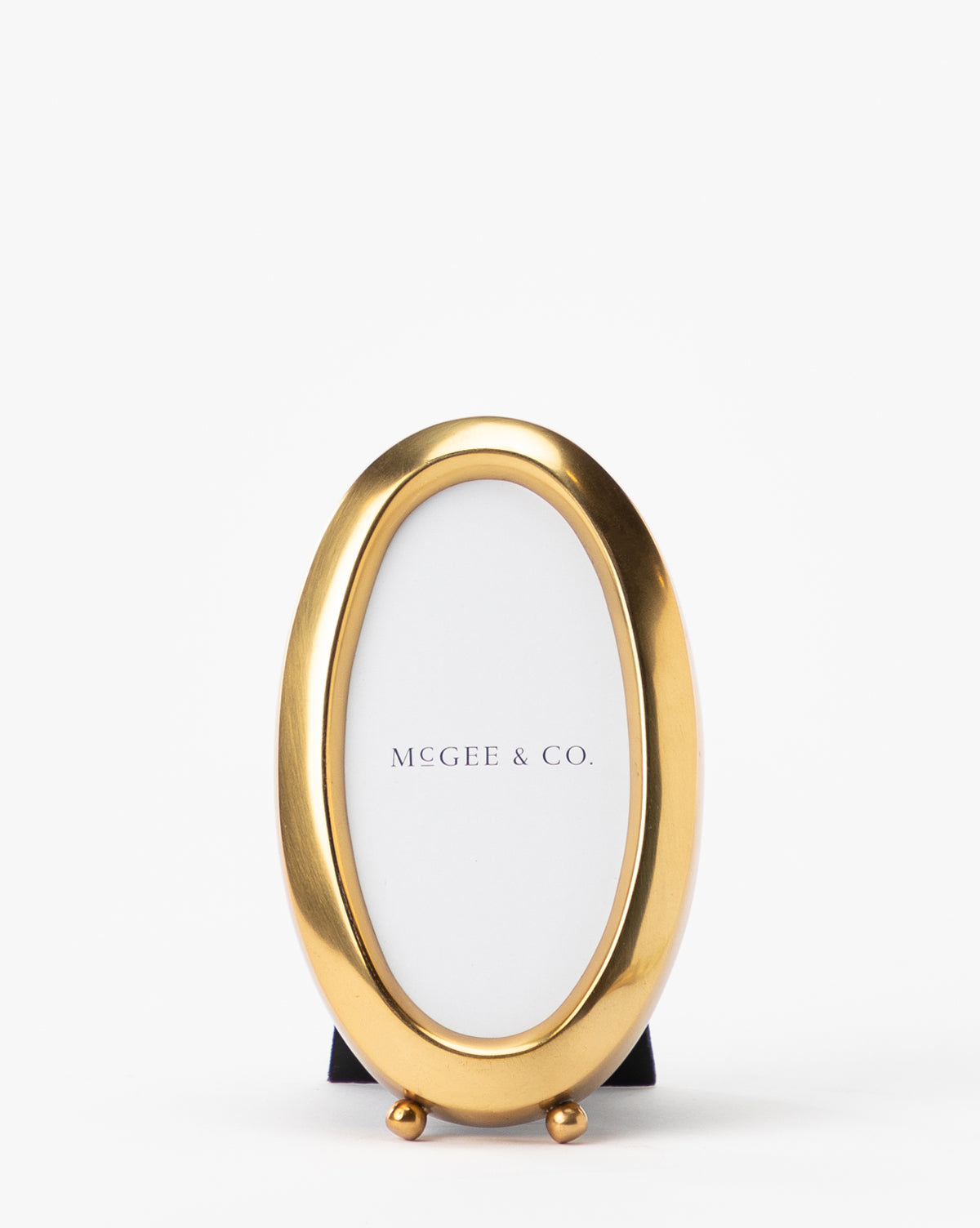 An oval Blanche Picture Frame with an antique brass, shiny finish stands upright against a white background, featuring McGee & Co. on the insert.