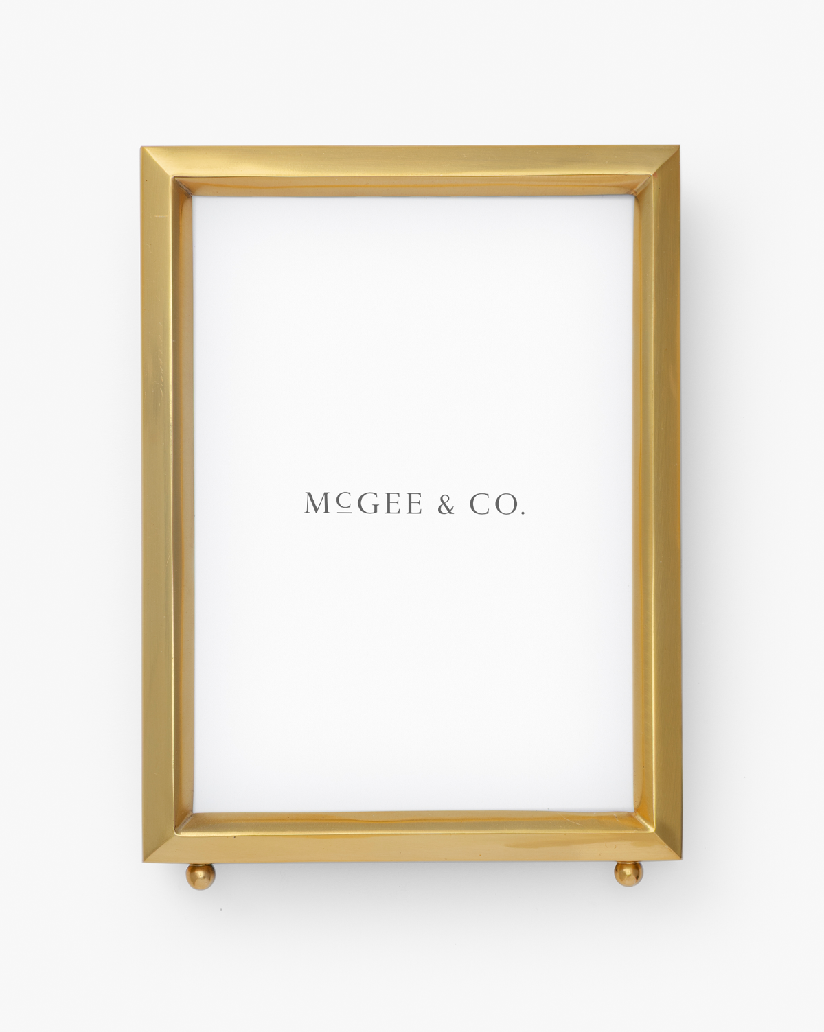 The Blanche Picture Frame is a rectangular gold frame with a white background, featuring "MCGEE & CO." centered. Its modern, elegant design sits on two small gold spheres at the base, by McGee & Co.