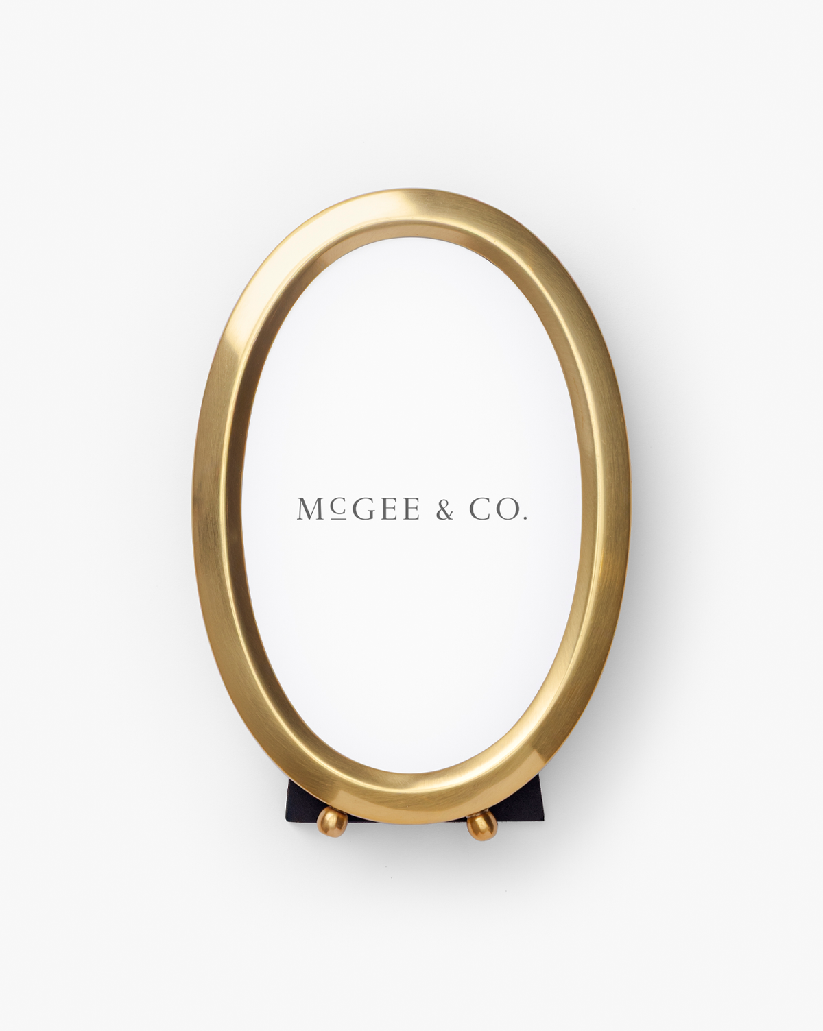 The Blanche Picture Frame is an oval, elegant photo frame with a polished brass finish, a white insert displaying "McGee & Co.," and a small black stand at the bottom, all shown on a plain white background, by McGee & Co.