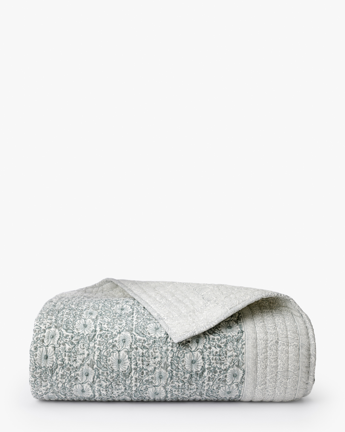 The Blue Floral Block Print Quilt, featuring a neatly folded design with a textured light gray and white pattern, is displayed on a plain white background. A turned-over corner reveals the artisanal bedding’s reverse side, by McGee & Co.