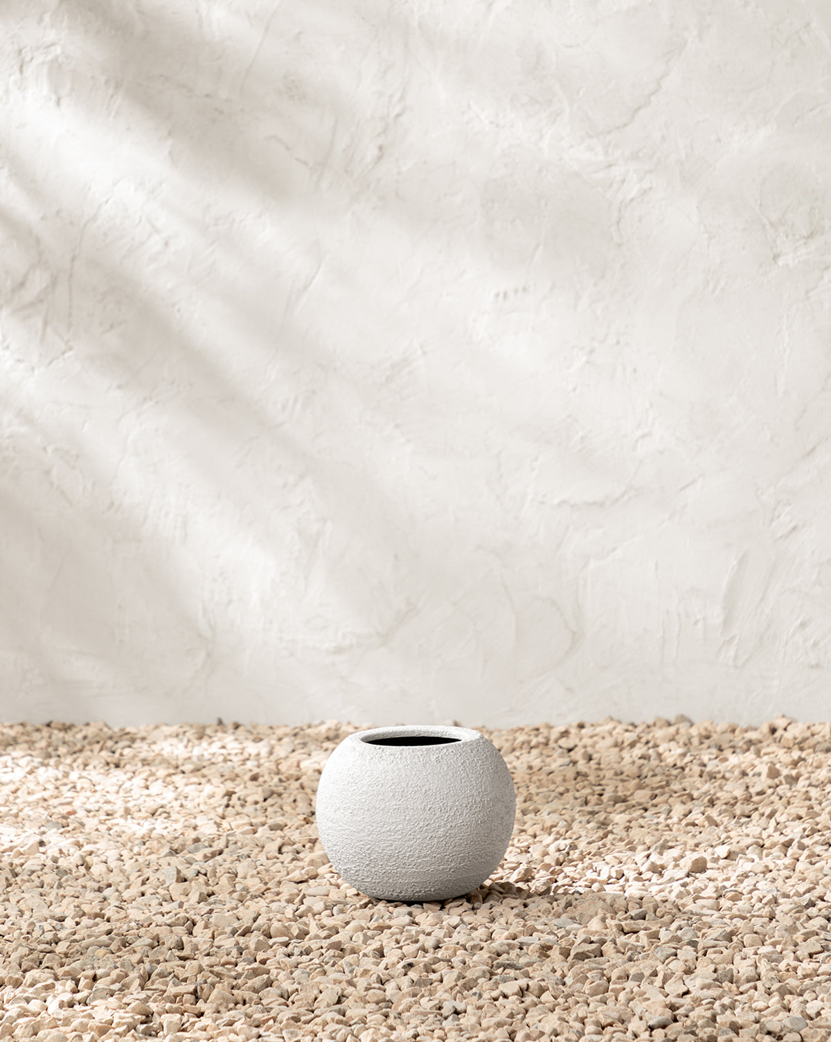 The Bodie Fiberstone Planter, featuring a round, textured white design, rests on beige pebbles against a softly lit, textured plaster wall.