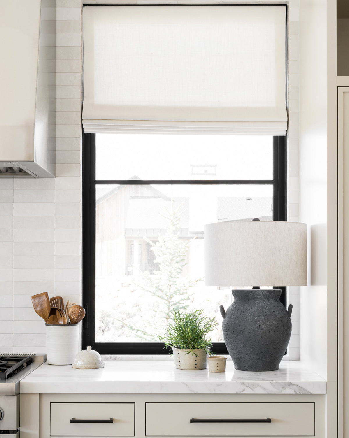 A bright kitchen features a marble countertop, terracotta potted plants, the Anza Table Lamp in gray, a white utensil holder with wooden utensils, and a window with a white roman shade letting in natural light, by McGee & Co.